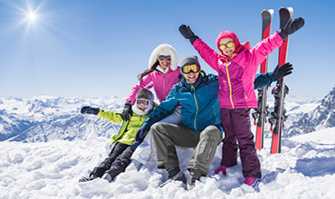 Eine vierköpfige Familie in farbenfroher Skiausrüstung und Skibrille posiert glücklich auf einem schneebedeckten Berg. Skistöcke und Skier stehen aufrecht im Schnee. Die strahlende Sonne scheint vom klaren blauen Himmel und hebt die schneebedeckten Gipfel im Hintergrund hervor.