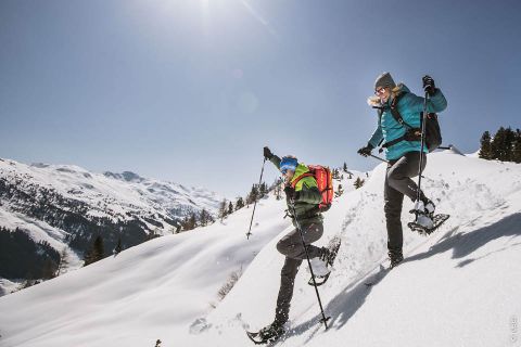 Zwei Personen wandern mit Schneeschuhen durch eine verschneite Berglandschaft unter strahlend blauem Himmel. Beide tragen Winterkleidung und Rucksäcke. Sie steigen einen Hang hinab, während die Sonne hell scheint und die schneebedeckten Gipfel im Hintergrund beleuchtet.