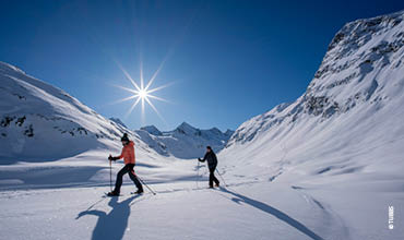 Zwei Personen fahren auf einem sonnenbeschienenen, schneebedeckten Berghang Ski. Über ihnen strahlt ein strahlend blauer Himmel und die Sonne scheint. Majestätische, schneebedeckte Gipfel umgeben die Skifahrer, während sie durch die unberührte Landschaft gleiten.