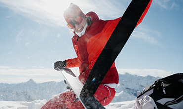 Eine Person in roter Skiausrüstung bewegt sich mit einem Skistock in der Hand auf einem verschneiten Hang. Die Sonne scheint hell vom klaren blauen Himmel. Im Hintergrund sind schneebedeckte Berge zu sehen. Die Szene vermittelt Energie und Freude an der Winterlandschaft.