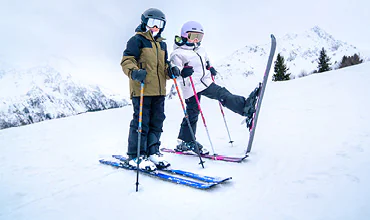 Zwei Kinder in Skiausrüstung stehen an einem verschneiten Hang. Eines hat spielerisch die Skier hochgehoben. Beide tragen Helme und Skibrillen. Im Hintergrund sind schneebedeckte Berge und Bäume zu sehen.