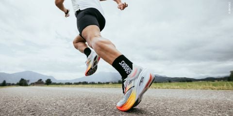 Eine Person in Sportkleidung läuft auf einer asphaltierten Straße, umgeben von Gras. Sie trägt bunte Laufschuhe und schwarze Socken. Der Himmel ist bewölkt.