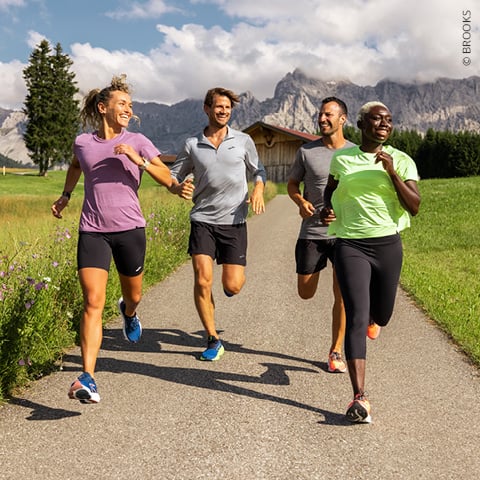 Vier Personen joggen energisch auf einem gepflasterten Weg durch eine malerische Landschaft mit Bergen, grünen Feldern und klarem Himmel im Hintergrund. Sie scheinen den Lauf zu genießen, lächeln und tragen lässige Sportkleidung.