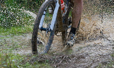 Eine Person fährt mit einem Mountainbike durch eine schlammige Pfütze und spritzt dabei Wasser und Schlamm auf. Das Bild zeigt eine Nahaufnahme der Fahrradreifen und der sich bewegenden Beine und unterstreicht die Dynamik des Radfahrens in unwegsamem Gelände.