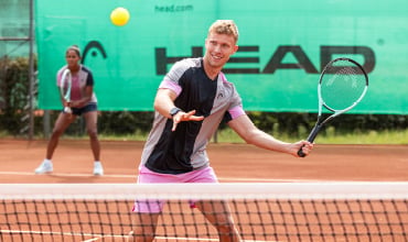 Ein Mann in schwarz-grauem Sportoutfit mit pinken Akzenten spielt Tennis auf einem Sandplatz und bereitet sich auf einen Vorhandschlag vor. Hinter ihm schaut eine Frau im passenden Outfit zu und ist bereit zum Spiel. Auf grünem Hintergrund ist ein großes „HEAD“-Logo zu sehen.