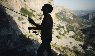 Silhouette einer Person, die in einer Berglandschaft klettert. Sie trägt einen Helm und hält ein Seil. Sie steht vor dem Hintergrund zerklüfteten Geländes und entfernter Hügel unter klarem Himmel.