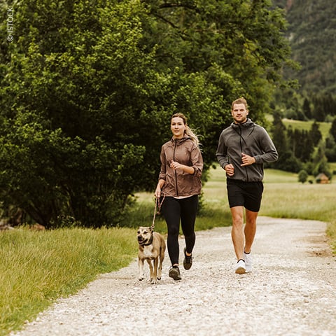 Ein Mann und eine Frau joggen Seite an Seite mit einem Hund an der Leine auf einem Schotterweg. Sie sind umgeben von saftigem grünem Gras und Bäumen, im Hintergrund sind Hügel zu sehen. Beide tragen Kapuzenpullover und Shorts und sind ganz auf ihre Aktivität konzentriert.