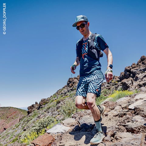 Ein Mann mit Mütze und Sonnenbrille läuft unter strahlend blauem Himmel auf einem steinigen Pfad. Er trägt ein blaues Hemd und karierte Shorts, einen Rucksack und eine Armbanduhr und ist von einer rauen Landschaft mit spärlicher Vegetation umgeben.