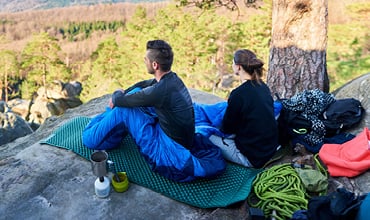 Zwei Personen in Wanderkleidung sitzen auf einer Isomatte auf einer Felsklippe und blicken auf eine Waldlandschaft. Neben ihnen liegen ein blauer Schlafsack, ein aufgerolltes grünes Seil und Campingausrüstung. Eine Person ist in den Schlafsack eingewickelt.
