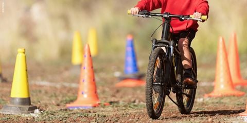 Eine Person in einem roten Kapuzenpullover fährt mit dem Fahrrad auf einem Feldweg eine mit gelben und orangefarbenen Verkehrskegeln markierte Strecke ab. Die Szene suggeriert Übung oder Training im Freien an einem sonnigen Ort.