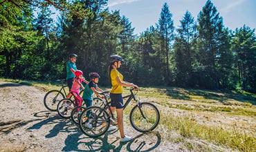 Eine vierköpfige Familie, darunter zwei Kinder, steht mit ihren Fahrrädern, Helmen und Fahrradausrüstung auf einem Feldweg in einem Waldgebiet. Im Hintergrund sind hohe Bäume und ein strahlend blauer Himmel zu sehen, die einen sonnigen Tag im Freien suggerieren.