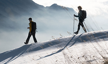 Zwei Wanderer wandern einen schneebedeckten Grat entlang. Einer benutzt Trekkingstöcke, der andere geht ohne Stöcke weiter. Berggipfel und Nebelwolken bilden den Hintergrund und schaffen eine ruhige und abenteuerliche Atmosphäre.