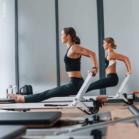 Zwei Frauen machen in einem Fitnessstudio Übungen an Pilates-Reformern. Beide tragen Sportkleidung und konzentrieren sich auf ihr Training. Der Raum hat große Fenster, die viel Tageslicht hereinlassen.