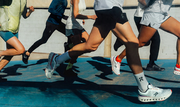 Eine Gruppe von Menschen läuft auf einer Laufbahn. Das Bild konzentriert sich auf ihre Beine und Füße in Sportschuhen. Sie bewegen sich und werfen Schatten auf die blaue Fläche. Einige tragen Shorts, im Hintergrund ist eine helle Wand zu sehen.