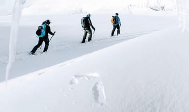 Drei Personen in Winterkleidung wandern durch eine verschneite Landschaft. Sie stehen in einer Reihe und laufen mit Wanderstöcken durch den tiefen Schnee. Die Szene ist hell und weiß und suggeriert eine kalte, winterliche Umgebung ohne sichtbare Vegetation.