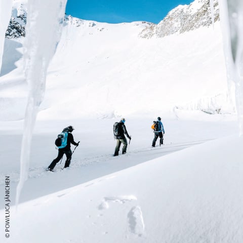 Drei Personen wandern durch eine verschneite Landschaft, umgeben von eisigen Klippen. Sie sind mit Winterausrüstung ausgestattet, darunter Rucksäcke und Trekkingstöcke. Der Himmel ist klar und blau und bildet einen Kontrast zum weißen Schnee.