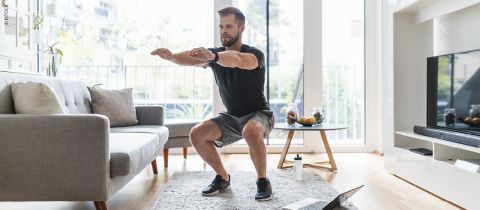 Mann macht Kniebeugen in einem Wohnzimmer mit grauem Sofa und Couchtisch. Er trägt ein schwarzes Hemd und graue Shorts. Auf dem Boden steht ein Laptop. Im Hintergrund ist ein großes Fenster mit Blick auf den Balkon zu sehen.