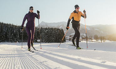Zwei Personen laufen unter klarem Himmel auf einer verschneiten Loipe Langlaufski. Sie tragen Winterkleidung, die eine in Lila, die andere in Gelb. Im Hintergrund sind schneebedeckte Bäume und Berge zu sehen.