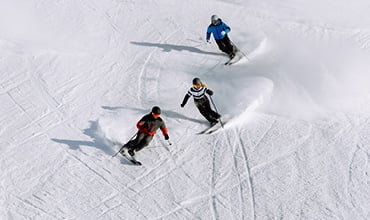 Drei Personen fahren einen schneebedeckten Hang hinunter. Sie tragen Winterkleidung in den Farben Rot, Blau und Schwarz und hinterlassen geschwungene Spuren. Die Sonne wirft Schatten auf den unberührten Schnee und unterstreicht ihre Bewegungen.