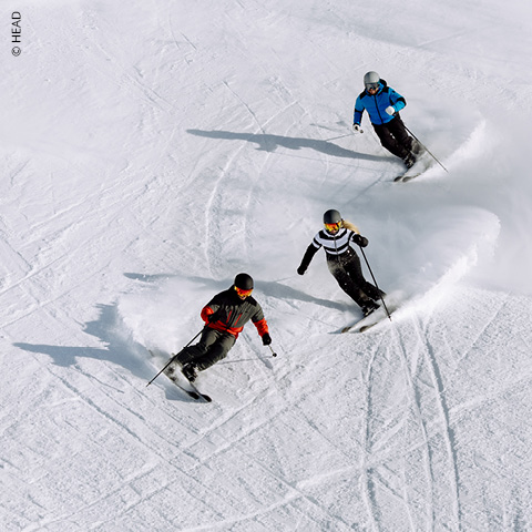 Drei Skifahrer fahren parallel zueinander einen verschneiten Hang hinunter. Sie tragen Winterkleidung und Helme und ziehen ihre Spuren durch den Schnee. Der strahlend weiße Schnee bildet einen Kontrast zu ihren farbenfrohen Outfits.