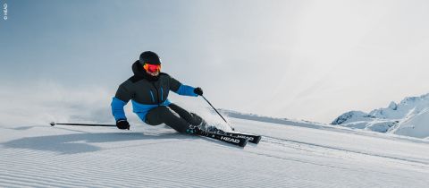 Ein Skifahrer in blau-schwarzem Outfit und roter Skibrille fährt an einem sonnigen, klaren Tag durch den Schnee, im Hintergrund sind Berggipfel zu sehen. Der Skifahrer legt sich in die Kurve und erzeugt so eine Schneespritze hinter sich.
