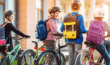Vier Kinder mit Helmen und Rucksäcken fahren an einem sonnigen Tag Fahrrad. Sie sind leger gekleidet und scheinen sich zu unterhalten. Sie fahren in der Nähe eines Gebäudes mit großen Fenstern.