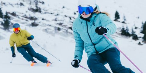 Zwei Personen fahren einen verschneiten Hang hinunter. Die Person im Vordergrund trägt eine blaugrüne Jacke und ein blaues Visier, die Person im Hintergrund eine gelbe Jacke und einen grauen Helm. Im Hintergrund sind schneebedeckte Bäume und Felsen zu sehen.