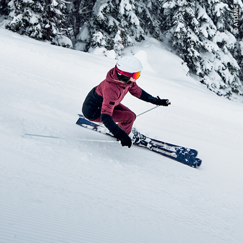 Ein Skifahrer in roter Jacke und weißem Helm fährt eine Kurve auf einem verschneiten Hang. Im Hintergrund sind schneebedeckte Bäume zu sehen, und in der Skibrille ist das Spiegelbild des Skifahrers zu sehen.