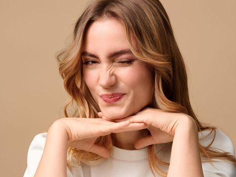 Eine Person mit hellbraunem Haar zwinkert verspielt und stützt ihr Kinn auf die Hände. Sie lächelt leicht und der Hintergrund ist in einem sanften Beige gehalten.