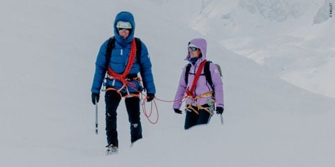 Zwei Personen in Winterkleidung wandern durch den Schnee. Sie tragen Helme und Klettergurte. Sie sind durch ein rotes Seil verbunden und tragen Eispickel. Im Hintergrund ist eine verschneite Berglandschaft zu sehen.