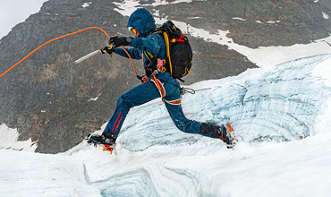 Ein Bergsteiger in blauer Ausrüstung, mit Helm und Eispickel, springt über eine schneebedeckte Gletscherspalte. Gesichert sind sie mit einem orangefarbenen Seil und Spikes an ihren Stiefeln. Im Hintergrund ist ein felsiger Berg zu sehen.