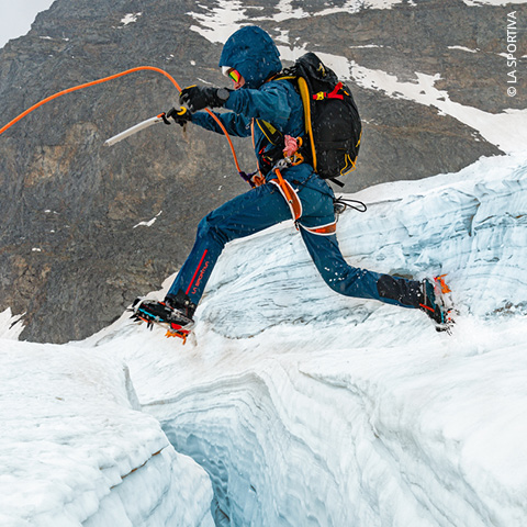 Ein Bergsteiger in blauer Ausrüstung und Steigeisen springt über eine Gletscherspalte auf einem schneebedeckten Gletscher. Er hält einen Eispickel und trägt einen Rucksack. Im Hintergrund sind felsiges Gelände und schneebedeckte Flächen zu sehen.