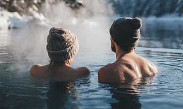 Zwei Menschen mit Strickmützen entspannen in einer dampfenden heißen Quelle, umgeben von verschneiten Landschaften. Sie sind teilweise im Wasser eingetaucht, mit dem Rücken zum Wasser, im Hintergrund sind Bäume und Schnee zu sehen.