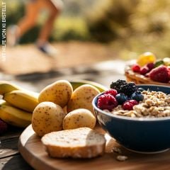 Ein Holztisch im Freien mit einem Teller Bananen, Kartoffeln und Beeren, einer Schüssel Müsli und Brot. Im unscharfen Hintergrund läuft eine Person auf einem Pfad. Sonnenlicht hebt die frischen Produkte hervor.