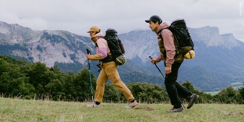 Zwei Personen mit Rucksäcken und Outdoor-Kleidung wandern mit Trekkingstöcken auf einem grasbewachsenen Hügel, im Hintergrund sind Berge und Bäume unter einem bewölkten Himmel zu sehen.
