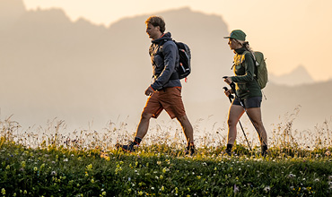 Zwei Menschen mit Rucksäcken und Outdoor-Kleidung wandern durch eine Wiese mit Wildblumen, im Hintergrund sind unter einem warmen, dunstigen Himmel sanft Berge zu erkennen.