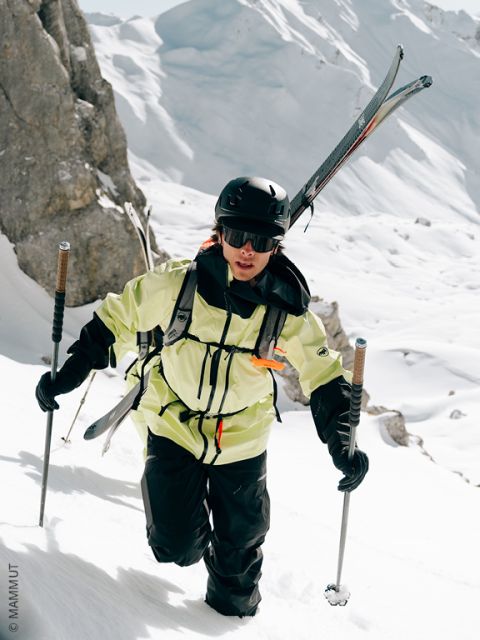 Ein Skifahrer mit Helm, Brille und gelber Jacke klettert einen verschneiten Berghang hinauf, wobei er die Skier auf dem Rücken trägt und die Skistöcke zur Unterstützung benutzt. Felsige Klippen und tiefer Schnee umgeben die Szene.