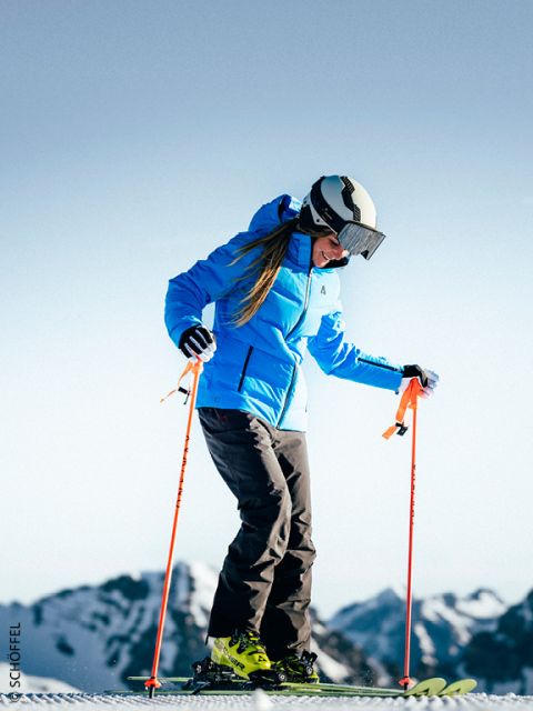 Person in blauer Jacke und Helm beim Skifahren auf Schnee mit Bergen im Hintergrund an einem klaren, sonnigen Tag.