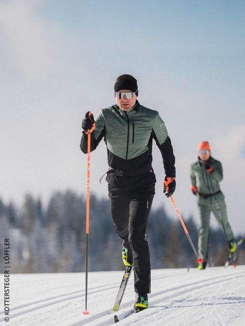 Zwei Skilangläufer auf einer verschneiten Loipe, mit Wintersportausrüstung und Sonnenbrille, mit Bäumen und blauem Himmel im Hintergrund.