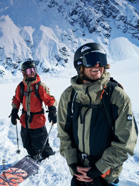 Zwei Snowboarder in Winterausrüstung stehen im Schnee, mit schneebedeckten Bergen im Rücken, lächeln und genießen den hellen, sonnigen Tag.