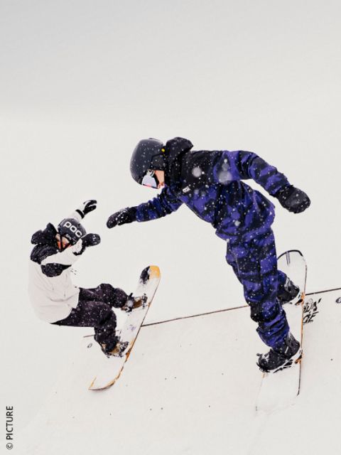 Zwei Snowboarder in Winterausrüstung führen Tricks auf einer verschneiten Rampe vor, umgeben von fallendem Schnee unter einem bedeckten Himmel.