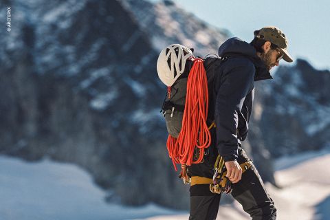 Ein Bergsteiger, der eine Ausrüstung trägt und einen Rucksack mit Helm und rotem Seil mit sich führt, geht in einer verschneiten Berglandschaft.