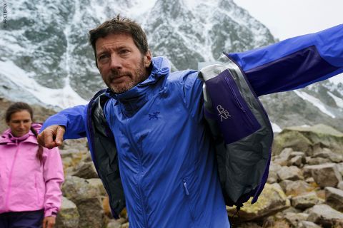 Ein Mann in einer blauen Jacke zieht beim Wandern in felsigen Bergen eine äußere Schicht an; eine Frau in einer rosa Jacke steht im Hintergrund.