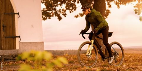 Ein Radfahrer in schwarzem Outfit und Helm fährt an einem sonnigen Tag eine kurvenreiche Bergstraße hinauf. Die Sonne wirft lange Schatten auf die Straße und im Hintergrund ist eine Felsklippe zu sehen.