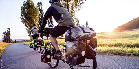 Ein Radfahrer fährt an einem klaren Tag Rennrad. Der Fahrer trägt einen Helm, eine Sonnenbrille und ein schwarzes Radsport-Outfit. Im Hintergrund ist eine Berglandschaft unter blauem Himmel zu sehen.
