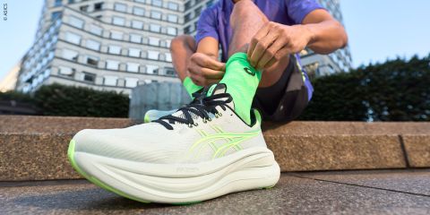 Im Freien sitzende Person, die hellgrüne Laufschuhe mit passenden Socken bindet, vor einem modernen Stadtgebäude.