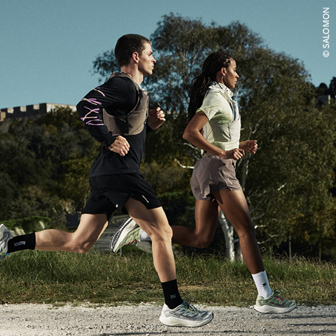 Zwei Personen laufen im Freien auf einem Kiesweg, umgeben von Bäumen, in Sportkleidung und -schuhen unter blauem Himmel.