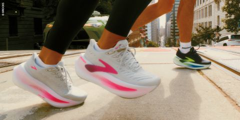 Zwei Personen laufen in weißen Laufschuhen mit leuchtend rosa und grünen Akzenten eine Stadtstraße hinauf, mit Gebäuden und einem Auto im Hintergrund.