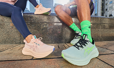 Zwei Personen in Sportkleidung sitzen im Freien und zeigen ihre schicken Laufschuhe - ein Paar ist blassrosa, das andere hellgrün mit neongrünen Socken.