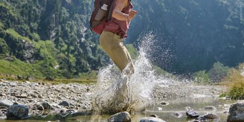 Eine Person mit einem Rucksack watet durch einen seichten Bach, umgeben von Felsen und Grün in einer bergigen Landschaft.